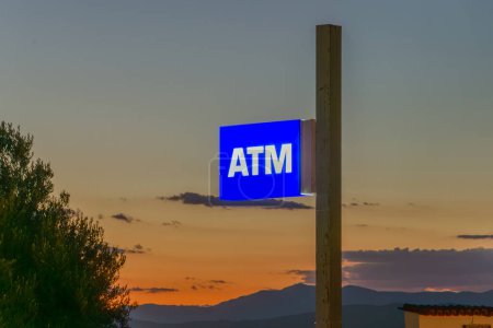 Photo for Glow-in-the-dark ATM sign. White letters on a blue background. The sky below is illuminated by the setting sun. - Royalty Free Image