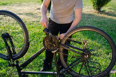 Photo for Man standing and repairing bicycle. Summer in green park. He is behind bicycle. repairman is wearing grey T-shirt and black trousers. - Royalty Free Image