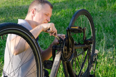 Photo for Man is repairing a bicycle chain in summer park. He is wearing a grey t-shirt. bicycle is positioned diagonally in the frame. surrounding space is green field. - Royalty Free Image