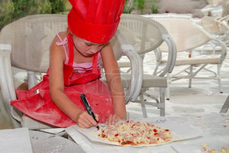 Photo for Five year old girl in red chef cap apron apron makes pizza. She is on summer terrace with white chairs and table - Royalty Free Image