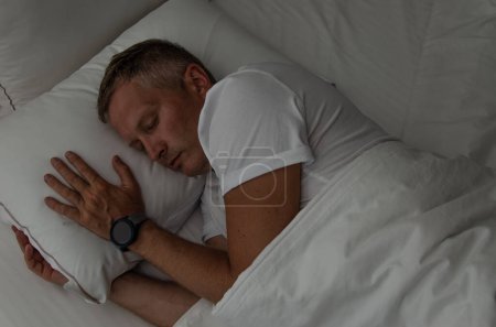 Foto de Reloj inteligente en el brazo del hombre dormido. Hombre blanco de mediana edad. Ropa interior blanca y almohada - Imagen libre de derechos