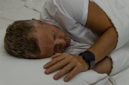 Foto de Reloj inteligente en el brazo del hombre dormido. Hombre blanco de mediana edad. Ropa interior blanca y almohada - Imagen libre de derechos