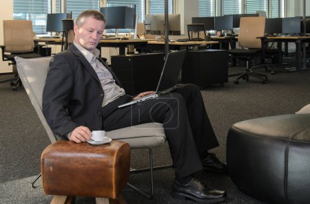 Foto de Empleado en el trabajo en la oficina de TI moderna. el hombre con el ordenador portátil y la taza de café se sienta en la silla en el área de reunión. Área de negocios de oficina en segundo plano. - Imagen libre de derechos
