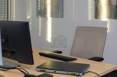 Photo for One workstation in IT office. There is monitor, keyboard, laptop on desk. A chair and glare of the sun in background. - Royalty Free Image