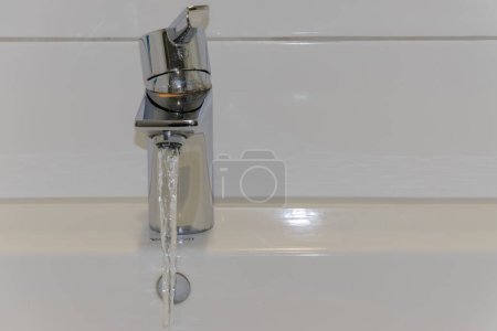 Photo for Close-up of water pouring from faucet into sink. White tiled wall and white ceramic sink. Background, copy space, text space - Royalty Free Image