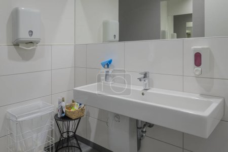 Photo for Pristine modern bathroom featuring long white sink, hand dryer, and soap dispenser mounted on tiled wall. A basket with cleaning supplies sits beside sink - Royalty Free Image