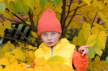 five-year-old girl next to an autumn yellow tree. She is wearing yellow vest, orange hat and orange hoodie . child is pointing at something.