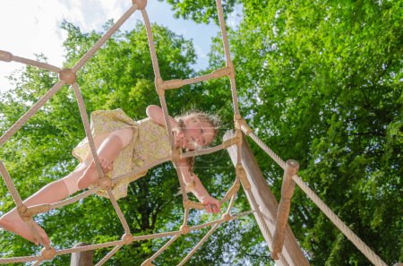 Photo for Five-year-old girl on climbing frame. She is blonde with two pigtails and barefoot. Greenery and sky in background. - Royalty Free Image