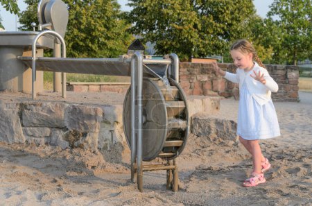 Photo for Child operating a manual water pump at a sandy playground. childs face is obscured. playground is surrounded by greenery, and there are trees in the background under a clear sky. - Royalty Free Image