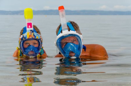 Photo for Child and an adult wearing snorkeling masks. Only their heads are visible above water. Faces in masks are reflected in water. Sea and sky are blue all around. Space for text. - Royalty Free Image