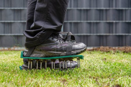 Nahaufnahme eines Rasenbelüftungsschuhs mit Metalldornen. Prozess der Bodenvertikutierung. Füße eines Mannes, der schwarze Schuhe trägt. Grünes Gras ringsum, anthrazitfarbener Zaun im Hintergrund.