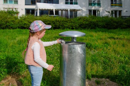 Girl throws trash into a street trash can. Trash can is metal, gray. Child of five with a long blonde braid. She is wearing a cap, pink vest and blue jeans. Theres greenery all around.