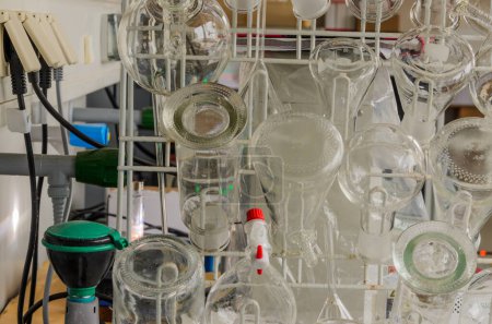 A metal rack in a laboratory holds various glass beakers in different shapes and sizes. Some beakers have markings indicating volume.
