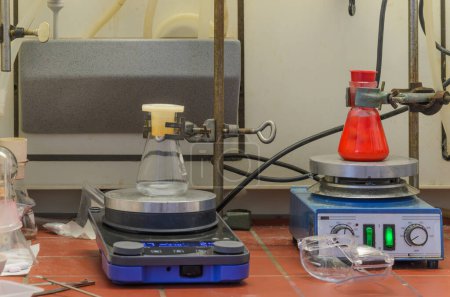 Laborgerät, das Magnetrührer mit weißer Rührstange und darauf platziertem Glasbecher enthält. Eine Flasche mit roter Flüssigkeit, die andere mit klarer Flüssigkeit.