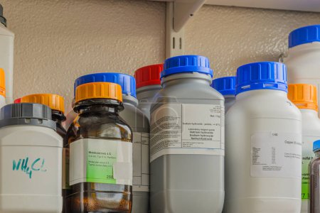 A shelf filled with various chemical bottles with labels. The labels contain information about the chemicals, including hazard symbols.