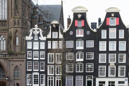 Façades brunes de bâtiments dans le vieil Amsterdam. Fenêtres blanches. Les toits à pignon sont décorés d'ornements. Une partie de la cathédrale est visible à gauche.