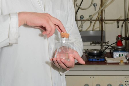A flask in the hands of a laboratory assistant close-up. The man is wearing a white protective robe, his hands are without gloves. In the background is a camera with a screwdriver.
