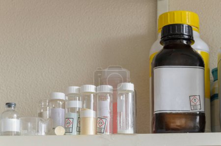 Une étagère remplie de diverses bouteilles chimiques avec des étiquettes. Les étiquettes contiennent des informations sur les produits chimiques, y compris les symboles de danger.