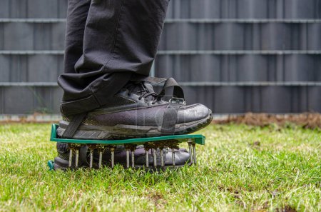 Nahaufnahme eines Rasenbelüftungsschuhs mit Metalldornen. Prozess der Bodenvertikutierung. Füße eines Mannes, der schwarze Schuhe trägt. Grünes Gras ringsum, anthrazitfarbener Zaun im Hintergrund.