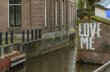 Photo for Brick wall along a canal in Amsterdam with the inscription Love Me. Letters are in white, bold, block letters. Brick building to the left is reflected in the water. - Royalty Free Image