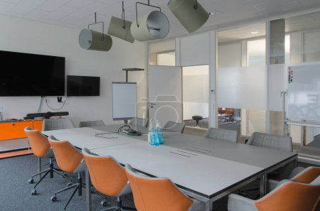 Photo for Well-lit conference room with long white table and several chairs set up for meeting. Flipchart and whiteboard and monitor tv screen are mounted on the wall at front of room. Lamps soffits on ceilling - Royalty Free Image