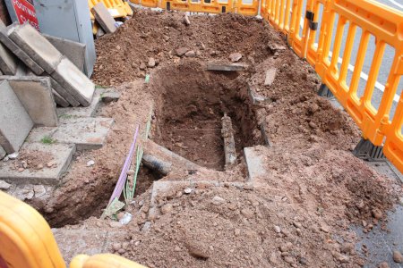 Road works, pavement dug up with exposed utility pipe and electrical cables and yellow plastic barrier, large hole in pavement