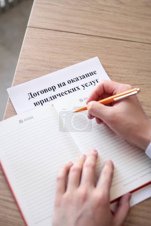 Foto de Tiro vertical mostrando un escritorio con un cuaderno abierto, acuerdo de servicio en ruso debajo, y las manos de un hombre, mano derecha en posición con un bolígrafo sobre el cuaderno en blanco. - Imagen libre de derechos