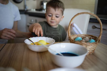 Photo for Little cute boy paints eggs for easter sitting in the kitchen at the table. - Royalty Free Image