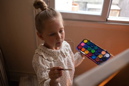 Photo for Little cute girl draws with paints on canvas at the easel. - Royalty Free Image