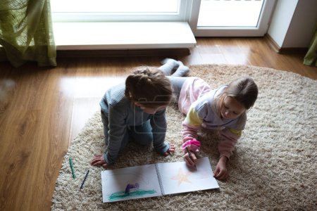 deux petites filles mignonnes dessinent dans un album couché sur le sol près d'une grande fenêtre.