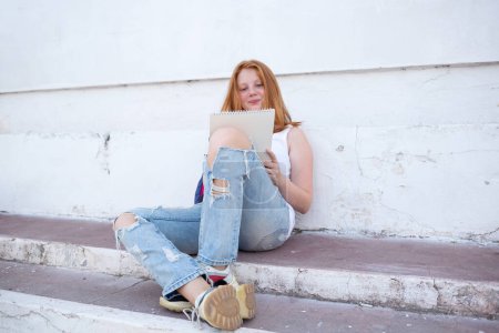Photo for Teenage girl draws in the sketchbook while sitting on the steps. - Royalty Free Image