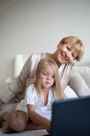 little girl with her grandmother sitting on the bed in front of a laptop.