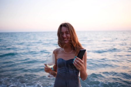 Foto de Hermosa joven con copa de vino y teléfono al atardecer junto al mar. - Imagen libre de derechos