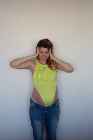 A joyful pregnant woman in a yellow bodysuit and jeans, smiling with excitement and anticipation.
