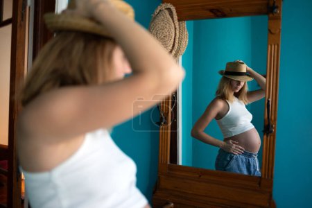 Pregnant woman in hat, smiling at her reflection in a mirror, blue wall background.