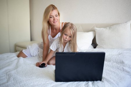 little girl with her mother sitting on the bed in front of a laptop.