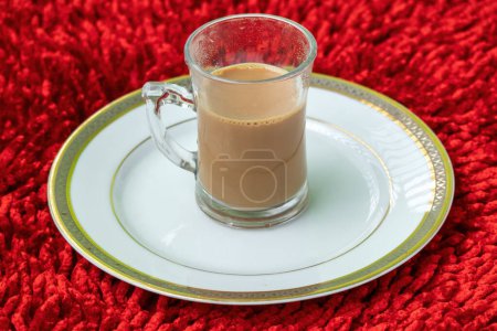 Una taza de té con leche caliente en un plato blanco sobre un fondo con textura de alfombra roja. Localmente en Bangladesh, se llama Dudh Cha. El té es la segunda bebida más popular del mundo.