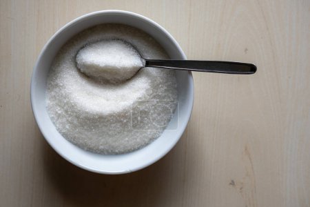 A white bowl and spoon full of sugar on a wooden background. Food ingredients for cooking sweets or desserts. Top view.