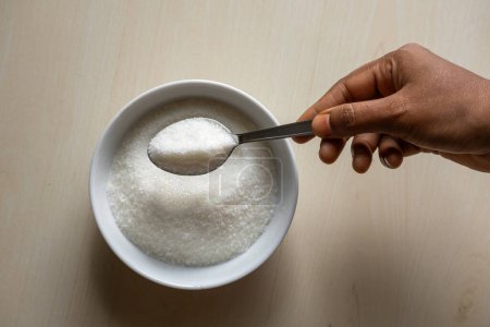 A white bowl is filled with sugar on wooden background. Woman's hand holding a sugar-filled spoon. Food ingredients for cooking sweets or desserts.