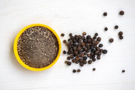 Crushed black pepper in a yellow container and whole black peppers isolated on a white background. Black pepper is a low-calorie spice that adds flavor to food without adding extra calories.