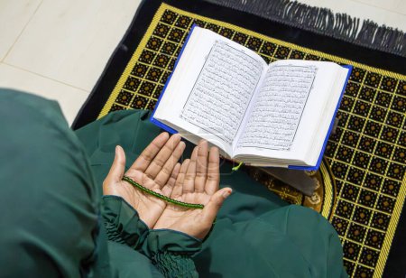 Junge muslimische Frau im Hijab, die "dua" (muslimisches Gebet) rezitiert, nachdem sie das heilige Buch Al Qur 'an rezitiert hat. Muslimische Frau betet mit grüner Tasbeeh in der Hand.