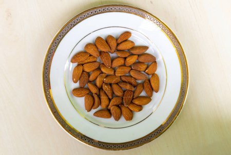 Top view of almond nuts on white plate on wooden background. Almonds are rich in nutrients, including fiber, magnesium, vitamin E, calcium, phosphorus, biotin, and monounsaturated fats. 