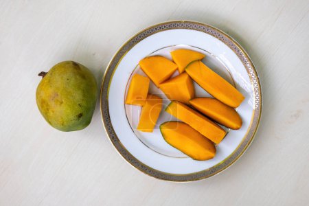 Peeled mango slices on a white plate on wooden background, a whole ripe mango placed beside. Top view
