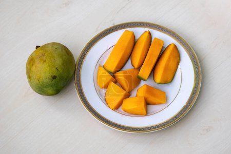 Peeled mango slices on a white plate on wooden background, a whole ripe mango placed beside