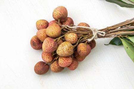 A bunch of fresh lychees tied together with a string, placed on a white wooden surface. The lychees have a reddish-brown textured skin.