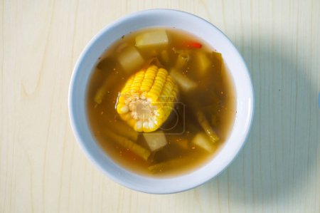 Indonesian traditional food named "sayur asem" with ingredient slices of chayote, corn, tamarind, long beans, chili pepper, bilimbi served on bowl isolated on wooden background