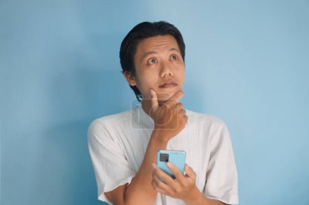 Photo for Young Asian man thinking about something while holding mobile phone - Royalty Free Image