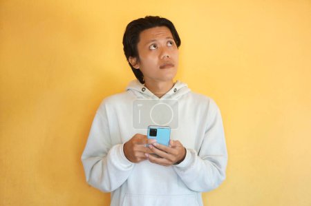 Photo for Asian young man wearing a hoodie thinking about something while holding mobile phone. - Royalty Free Image
