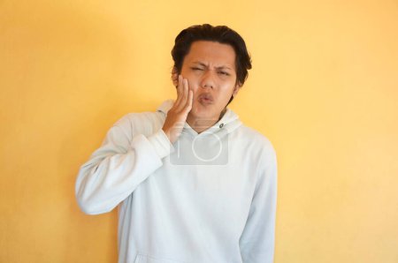 Asiatischer junger Mann trägt Kapuzenpullover mit Zahnschmerzen-Gesichtsausdruck.