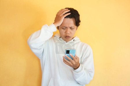 Asian young man showing confused facial expression while holding mobile phone